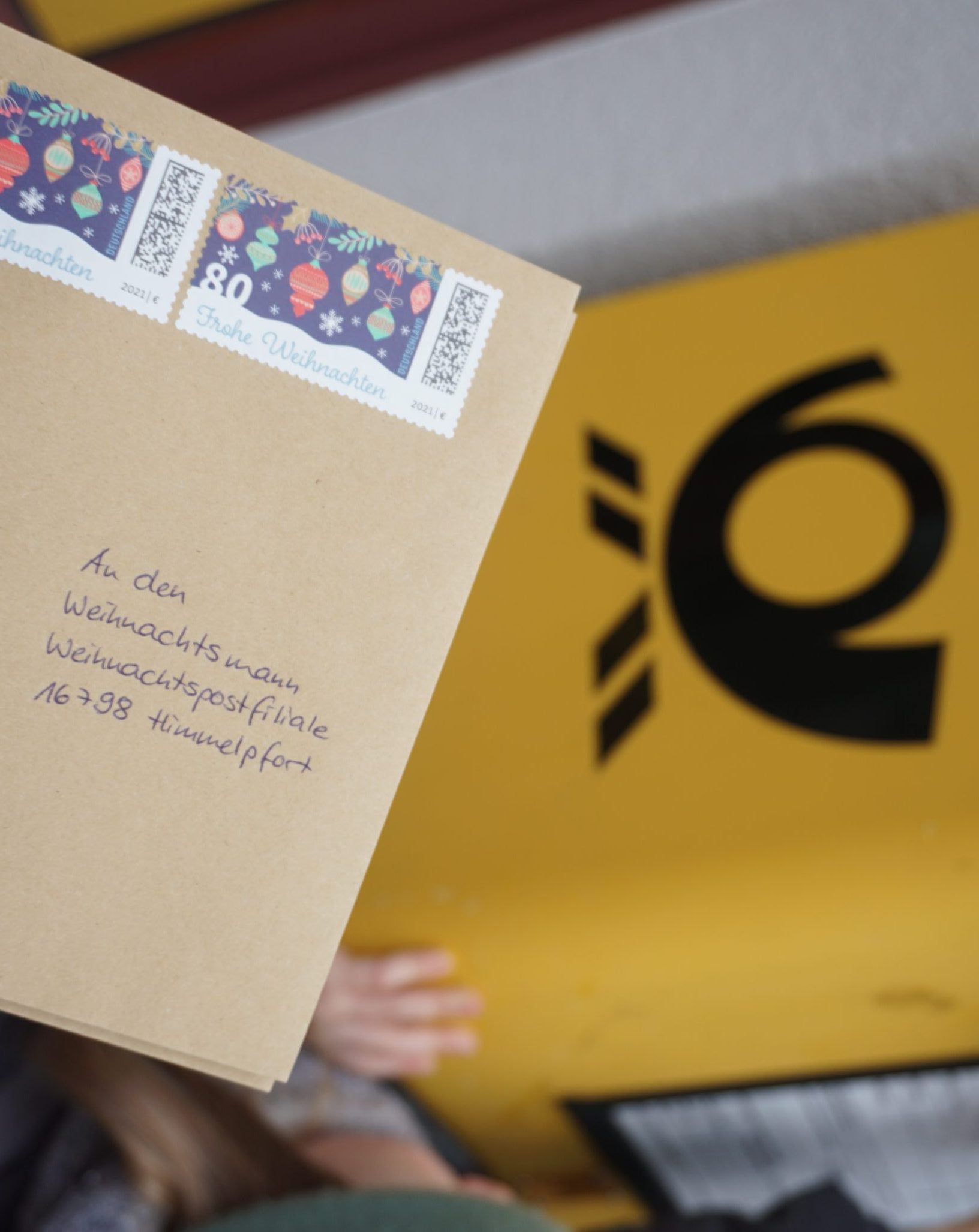Eine Hand hält einen Brief mit weihnachtlichen Briefmarken, der in deutscher Sprache an das Postamt Weihnachtsmann adressiert ist. So entsteht ein charmantes LebensBild vor einem gelben Briefkasten mit dem schwarzen Postsymbol. LebensBild