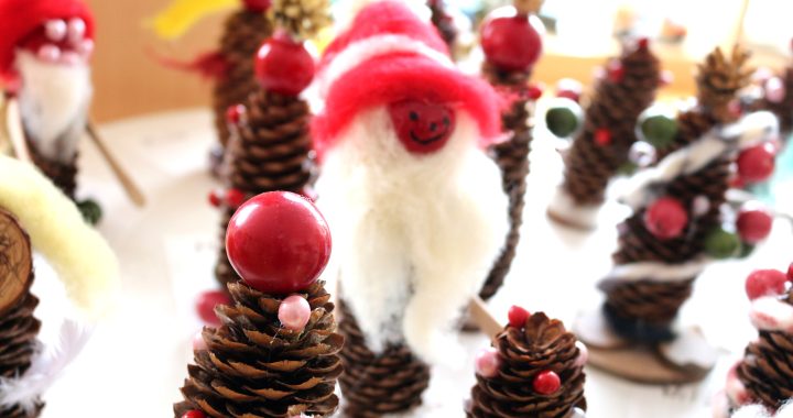 Nahaufnahme von Tannenzapfen, die mit rotem und weißem Filz, Perlen und Baumwolle dekoriert sind, um festlichen Weihnachtszwergen und Weihnachtsbäumen zu ähneln, angeordnet auf einem Tisch in hellem Tageslicht. LebensBild