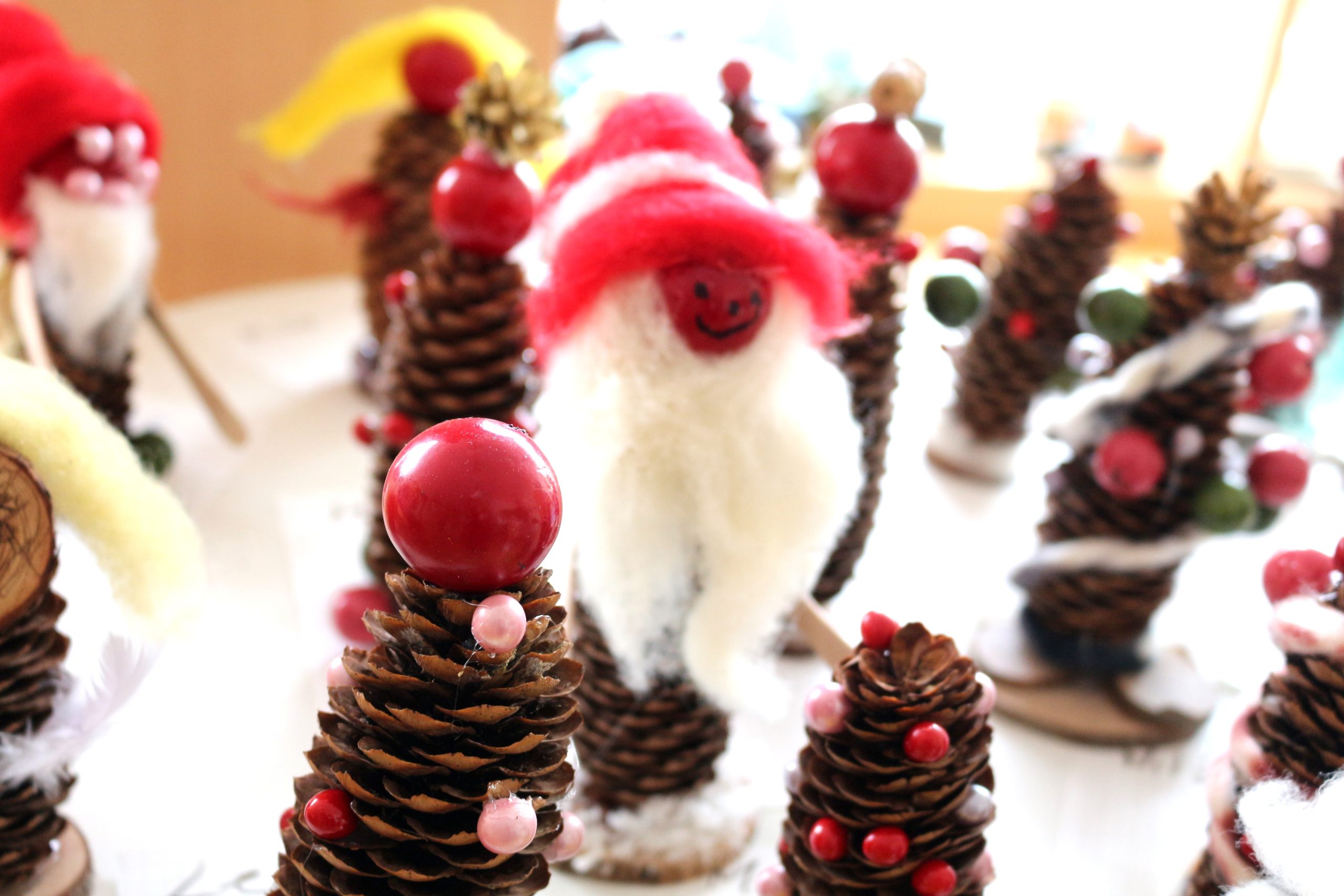 Nahaufnahme von Tannenzapfen, die mit rotem und weißem Filz, Perlen und Baumwolle dekoriert sind, um festlichen Weihnachtszwergen und Weihnachtsbäumen zu ähneln, angeordnet auf einem Tisch in hellem Tageslicht. LebensBild