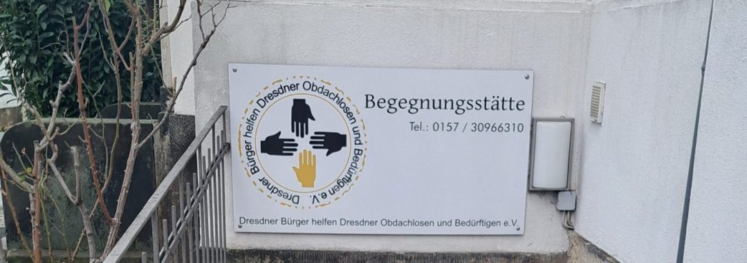 Eine Treppe führt hinunter zu einem Kellereingang eines Gebäudes. Über der Tür hängt ein Schild mit der Aufschrift Begegnungsstätte, einem Logo und Kontaktinformationen. Ein Besen und eine Kehrschaufel stehen neben der Tür; der Bereich ist sauber und ruhig.