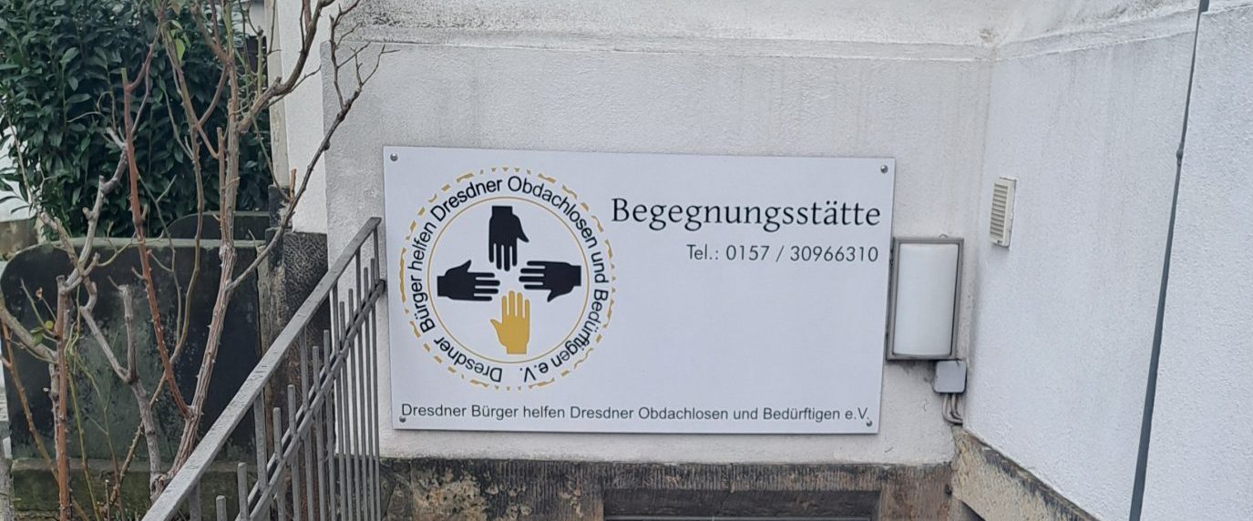 Eine Treppe führt hinunter zu einem Kellereingang eines Gebäudes. Über der Tür hängt ein Schild mit der Aufschrift Begegnungsstätte, einem Logo und Kontaktinformationen. Ein Besen und eine Kehrschaufel stehen neben der Tür; der Bereich ist sauber und ruhig.
