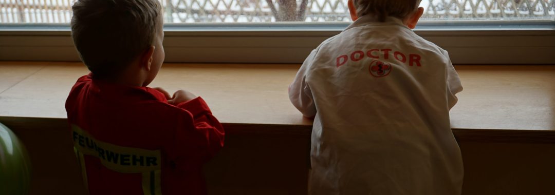 Zwei kleine Kinder stehen drinnen an einem Fenster und blicken auf einen verschneiten Hof hinaus.