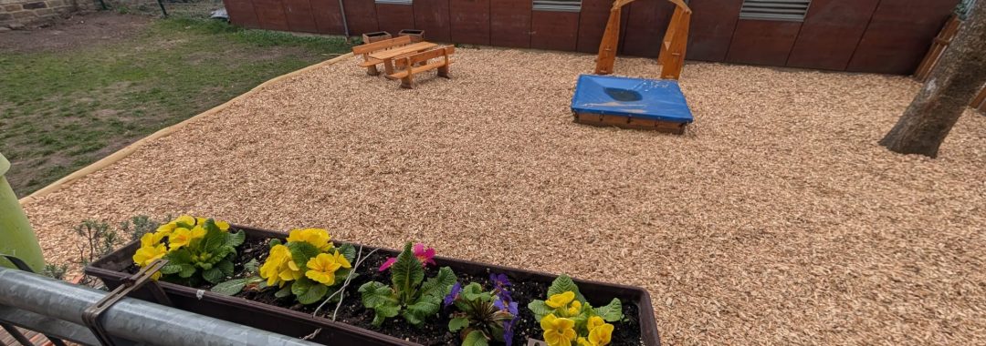 Ein Blumenkasten mit bunten Blüten ist an einem Balkongeländer aus Metall befestigt und überblickt einen Spielplatz mit Hackschnitzelboden, einem Sandkasten, einem Klettergerüst aus Holz und Picknicktischen. Im Hintergrund sind ein Baum und Gebäude zu sehen. LebensBild