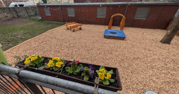 Ein Blumenkasten mit bunten Blüten ist an einem Balkongeländer aus Metall befestigt und überblickt einen Spielplatz mit Hackschnitzelboden, einem Sandkasten, einem Klettergerüst aus Holz und Picknicktischen. Im Hintergrund sind ein Baum und Gebäude zu sehen. LebensBild