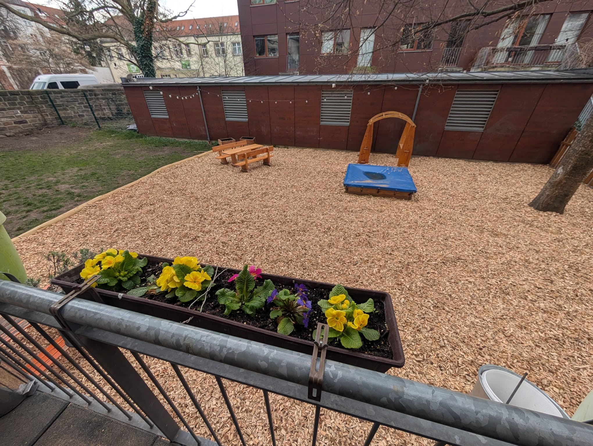 Ein Blumenkasten mit bunten Blüten ist an einem Balkongeländer aus Metall befestigt und überblickt einen Spielplatz mit Hackschnitzelboden, einem Sandkasten, einem Klettergerüst aus Holz und Picknicktischen. Im Hintergrund sind ein Baum und Gebäude zu sehen. LebensBild