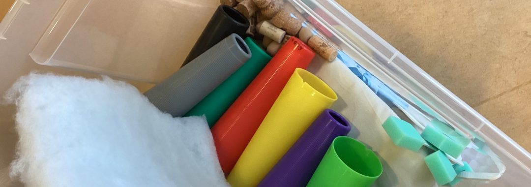 Ein Plastikbehälter enthält bunte Plastikröhren, grüne Schaumstoffblöcke, weiße Polster und einige Holzpflöcke, die alle lose im Inneren angeordnet sind. Der Behälter steht auf einer beigen Fläche.