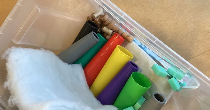 Ein Plastikbehälter enthält bunte Plastikröhren, grüne Schaumstoffblöcke, weiße Polster und einige Holzpflöcke, die alle lose im Inneren angeordnet sind. Der Behälter steht auf einer beigen Fläche.