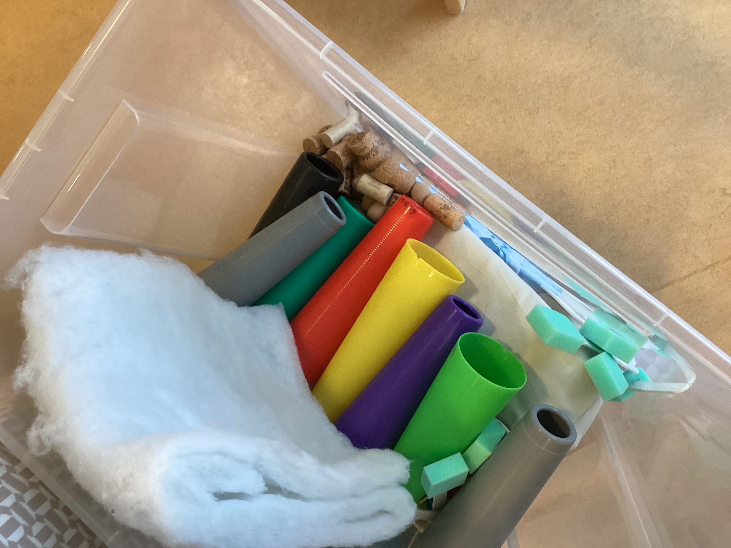 Ein Plastikbehälter enthält bunte Plastikröhren, grüne Schaumstoffblöcke, weiße Polster und einige Holzpflöcke, die alle lose im Inneren angeordnet sind. Der Behälter steht auf einer beigen Fläche.