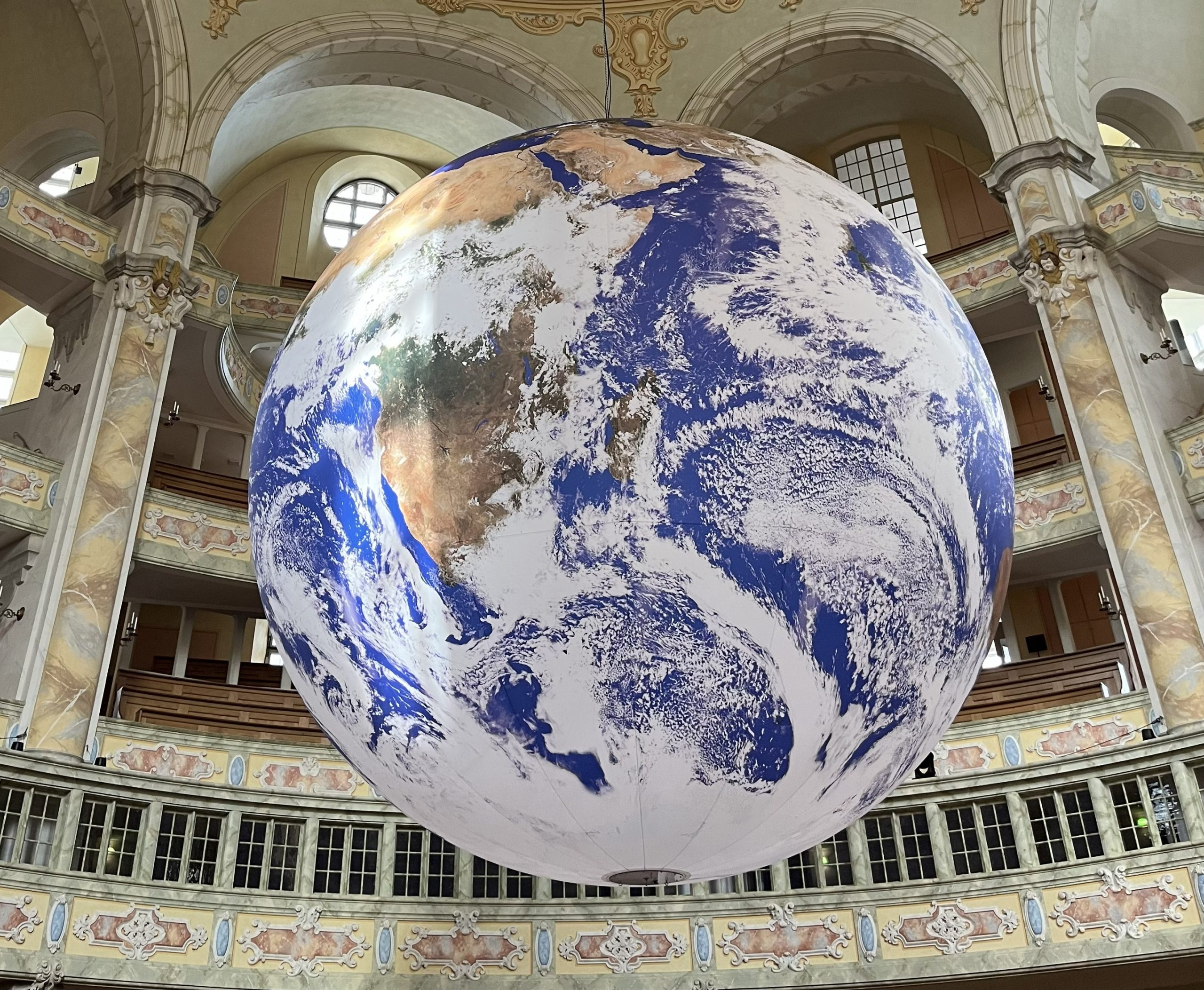 Ein großer, detaillierter Globus, der die Erde abbildet, hängt in einem prächtigen, verzierten Gebäude mit Bögen, Säulen und aufwändigen Verzierungen. Auf dem Globus sind vor allem Nord- und Südamerika abgebildet.