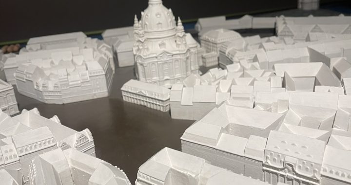 Ein detailliertes weißes Architekturmodell einer Stadtlandschaft mit einem markanten Kuppelgebäude im Zentrum, umgeben von verschiedenen geometrischen Strukturen, die Stadtblöcke und Straßen darstellen.