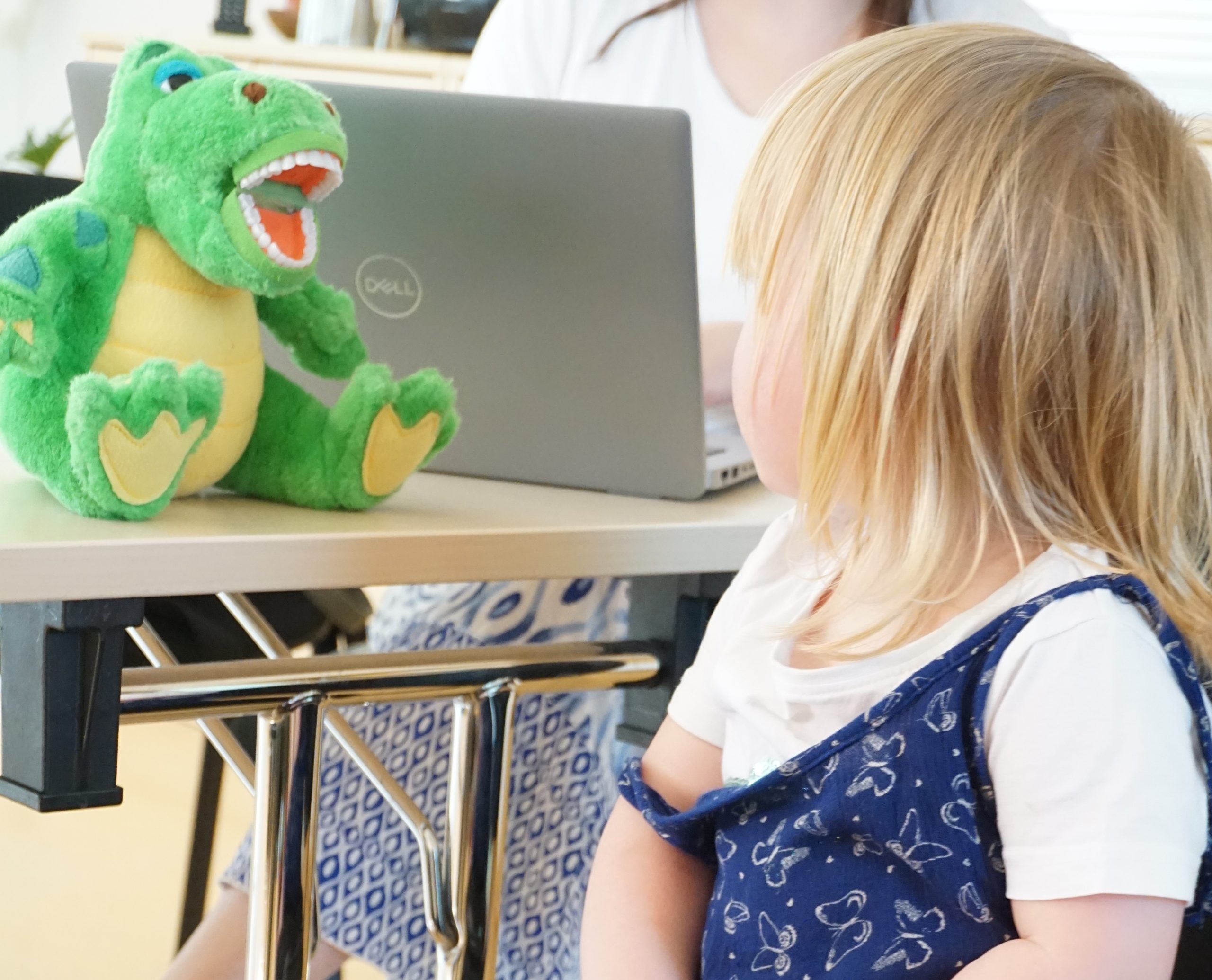 Ein kleines Kind mit blondem Haar betrachtet ein grünes Dinosaurier-Plüschtier, das auf einem Tisch sitzt. Hinter dem Tisch arbeitet eine Person mit einem grauen Dell-Laptop. Die Szene ist hell beleuchtet und befindet sich in einem Innenraum.