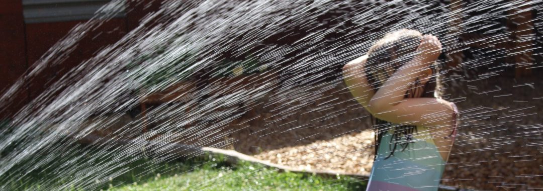 Ein Kind in einem bunten Badeanzug steht draußen und hält sich den Kopf, während es an einem sonnigen Tag unter dem Wasserstrahl eines Gartenschlauchs spielt. Die Wasserstrahlen sorgen im Vordergrund für einen funkelnden Effekt.