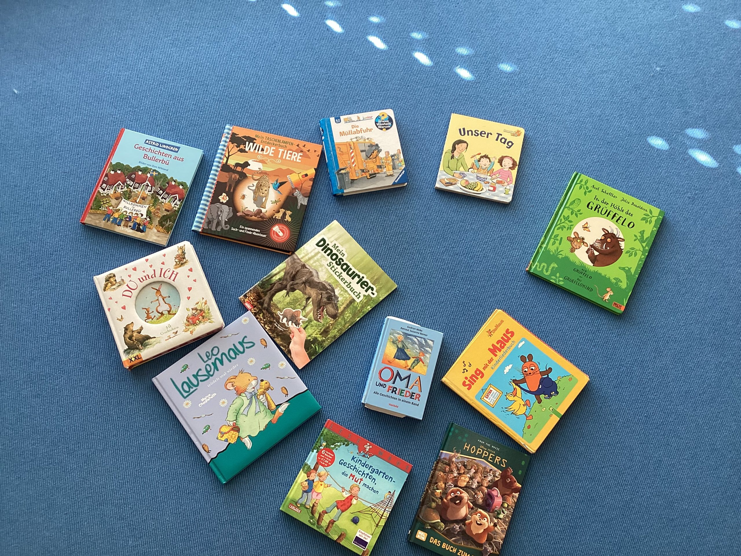 Dreizehn illustrierte deutsche Kinderbücher sind auf einem blauen Teppich in einer lockeren Herzform angeordnet. Die Bücher haben bunte Einbände und zeigen Tiere, Dinosaurier sowie Familienthemen.