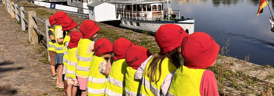 Eine Gruppe von Kindern in gelben Westen und roten Mützen steht an einem Geländer am Flussufer und beobachtet unter blauem Himmel die Boote auf dem Wasser, während im Hintergrund historische Gebäude zu sehen sind.