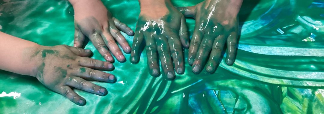 Drei mit grüner Farbe verschmierte Kinderhände verteilen und vermischen die Farbe auf einer ebenen Fläche und lassen so Wirbel und Muster entstehen.