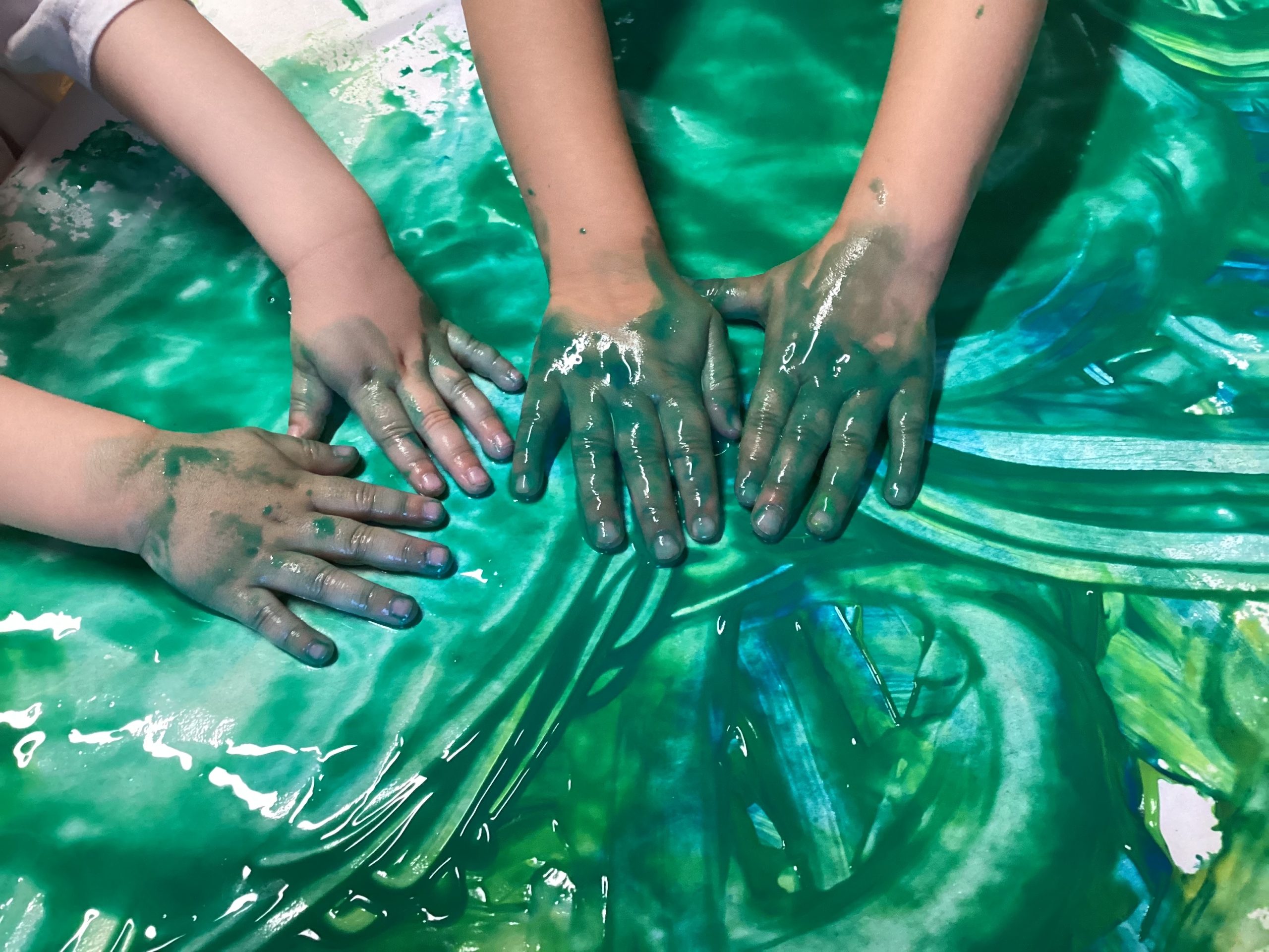 Drei mit grüner Farbe verschmierte Kinderhände verteilen und vermischen die Farbe auf einer ebenen Fläche und lassen so Wirbel und Muster entstehen.