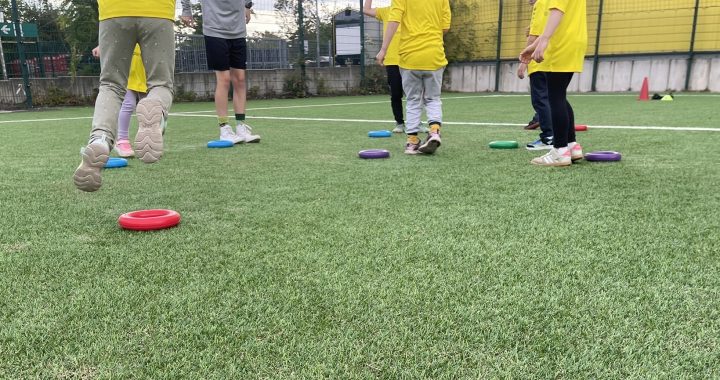 Kinder in Sportkleidung spielen auf einer Wiese und laufen zwischen bunten Ringen hin und her, die auf dem Boden verstreut liegen. Ein Trainer steht in der Nähe. Die Szene lässt auf eine Aktivität im Freien oder eine Trainingseinheit schließen.
