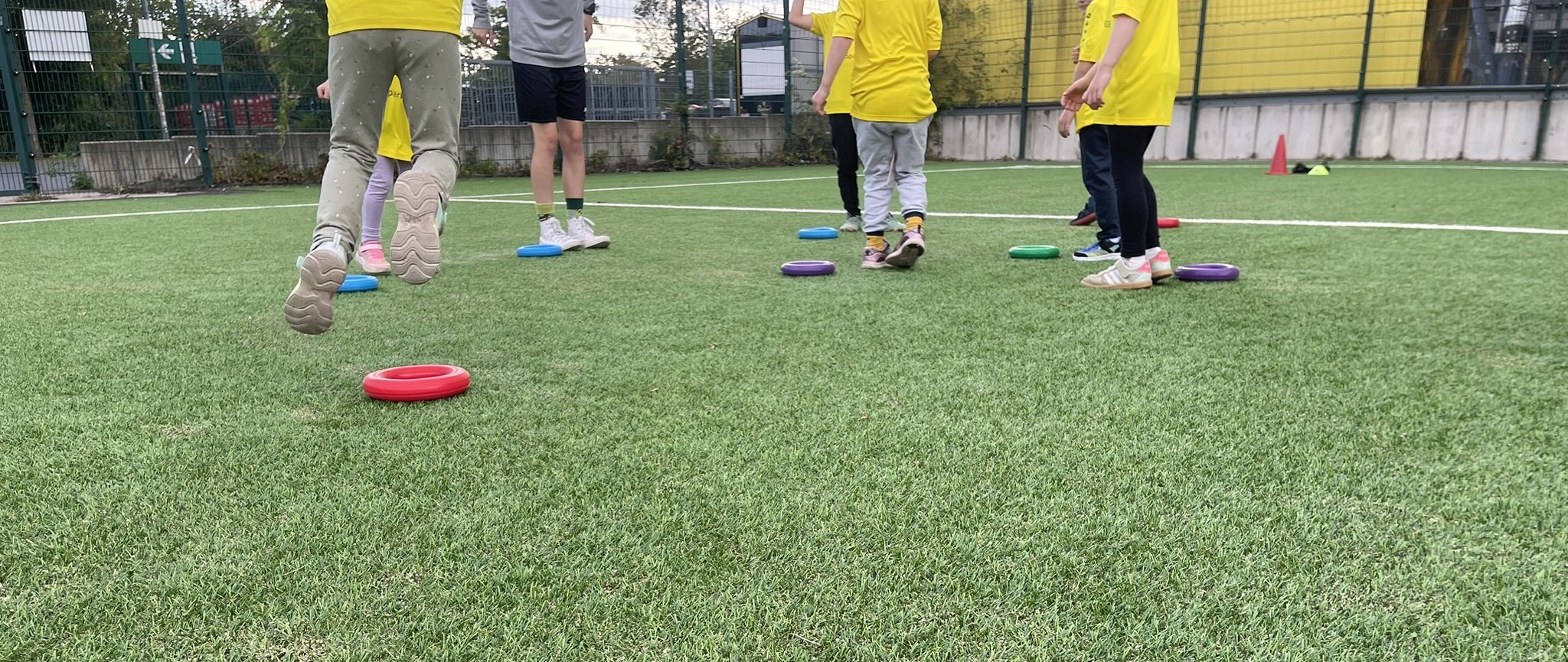Kinder in Sportkleidung spielen auf einer Wiese und laufen zwischen bunten Ringen hin und her, die auf dem Boden verstreut liegen. Ein Trainer steht in der Nähe. Die Szene lässt auf eine Aktivität im Freien oder eine Trainingseinheit schließen.
