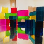 Bunte, durchsichtige Plastikspielzeugblöcke, wie die von Kita Lillabo, werden auf einer reflektierenden Oberfläche gestapelt und angeordnet. Ihre leuchtenden Gelb-, Rosa-, Grün- und Blautöne erzeugen geometrische Muster mit auffälligen Reflexionen.