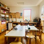 Ein helles, organisiertes Klassenzimmer oder ein Kunstraum - wie in der Kita Lillabo - mit Holzregalen, Tischen und Stühlen. Malutensilien, Papierrollen und Bastelmaterial sind übersichtlich angeordnet. Große Fenster füllen den Raum mit natürlichem Licht.