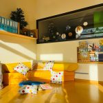 Eine gemütliche Leseecke in der Kita Lillabo besteht aus einem gelben Sofa, gepunkteten Kissen, einem Korb, Kinderbüchern in einem Wandregal und Sonnenlicht, das durch ein Fenster mit Blick in einen anderen Raum fällt.