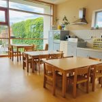 Ein helles, aufgeräumtes Klassenzimmer oder ein Essbereich in der Kita Lillabo ist mit Holztischen und -stühlen, großen Fenstern, die den Raum mit natürlichem Licht füllen, Küchengeräten an der Rückwand und üppigem grünen Efeu ausgestattet.
