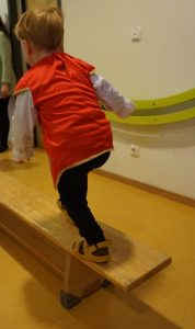 Ein kleines Kind in roter Weste und hellbraunen Sandalen balanciert auf einem Holzbalken in einem Innenraum, einen Fuß nach vorne und die Arme zum Ausgleich ausgestreckt. Eine Person steht im Hintergrund in der Nähe einer Türöffnung.