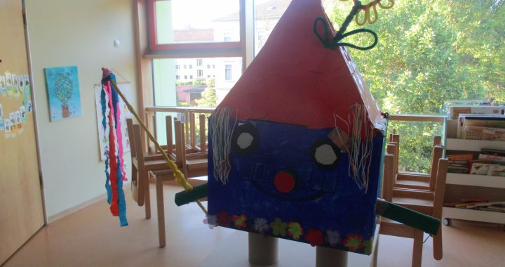 In einem hellen Gruppenzimmer mit großen Fenstern und Holzstühlen steht auf einem Tisch ein buntes Papphaus mit rotem Dach, das mit Blumen, einer Papierblume auf dem Dach und Pfeifenputzerarmen verziert ist.
