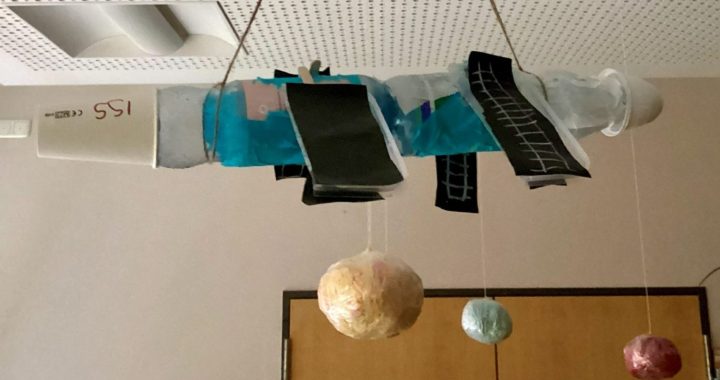 An einer perforierten Decke hängt ein handgefertigtes Mobile, bestehend aus einem verzierten horizontalen Rohr, an dem farbige Objekte befestigt sind, sowie Planeten, die an Schnüren darunter schweben. Im Hintergrund sind eine Holztür und beigefarbene Wände zu sehen.