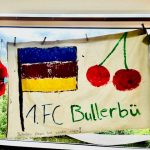 An einem Fenster hängt ein selbstgemachtes Banner mit einer gemalten Flagge, zwei roten Kirschen, der Aufschrift „1. FC Bullerbü“ und kleinen Bienenverzierungen am Fenster. Draußen sind Bäume und Gebäude zu sehen.