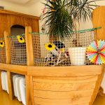 Eine große Spielanlage aus Holz in Form eines Schiffes mit Netz, Sonnenblumen-Dekorationen, bunten Papierfächern und einer Topfpflanze, aufgestellt in einem Innenraum mit heller Beleuchtung und festlicher Girlande.