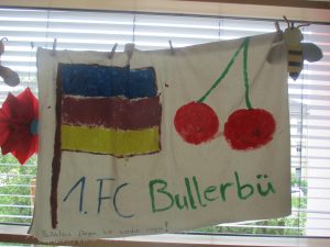 Auf einem selbstgemachten Banner sind eine Flagge mit blauen, roten und gelben Streifen, zwei Kirschen sowie der Schriftzug „1. FC Bullerbü“ in Grün und Blau zu sehen. Im Hintergrund sind Dekorationen und ein Fenster mit Jalousien zu erkennen.