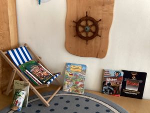 Eine kleine Leseecke mit einem gestreiften Liegestuhl, auf dem Kinderbücher liegen, einem Steuerrad an der Wand und weiteren Büchern, die auf einem blau-grauen Teppich ausgelegt sind. Eine maritime und gemütliche Atmosphäre.