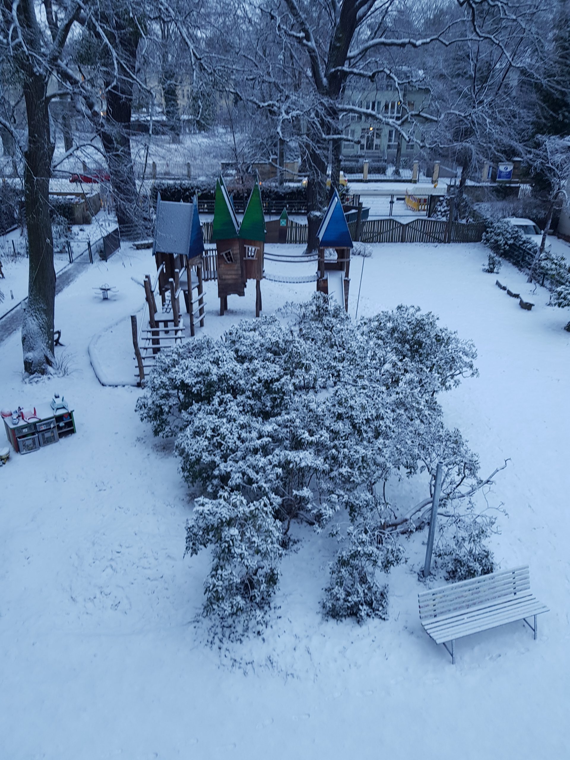 Ein schneebedeckter Spielplatz mit drei kleinen Kindervilla-Spielhäusern, einer Rutsche und einer Bank. Bäume und Sträucher sind mit Schnee bedeckt, und das Gelände ist von einem Holzzaun umgeben. Die Szene sieht ruhig und winterlich aus.
