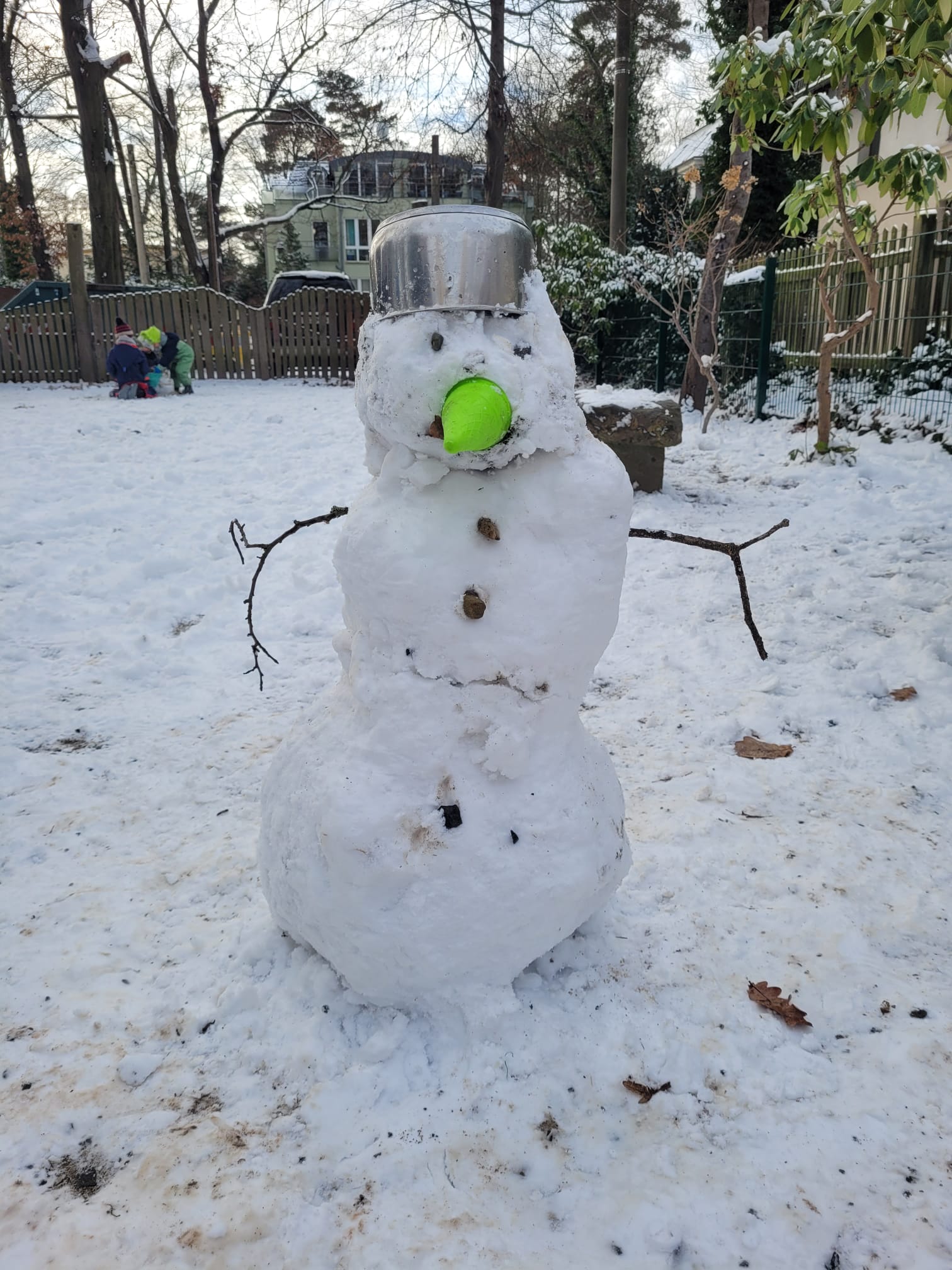 Ein Schneemann steht draußen im Schnee in der Kindervilla. Er trägt einen Topf als Hut, einen grünen Ball als Nase und Stöcke als Arme. Im Hintergrund sind Bäume, ein Zaun und fröhlich spielende Kinder zu sehen.