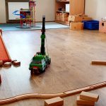 Ein bezauberndes Kindervilla-Spielzimmer mit Holzböden, verstreuten Holzklötzen, einer Holzeisenbahn, einem grünen Spielzeugkran und verschiedenen bunten Spielsachen und Aufbewahrungsboxen an den Wänden.