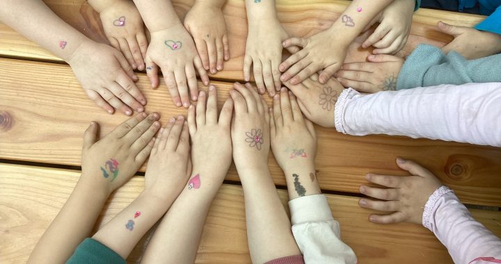 Kinderhände mit bunten temporären Tattoos, wie Blumen und Herzen, versammeln sich in einem Kreis auf einem Holztisch in der Kindervilla. Die Hände zeigen Ärmel in Blau, Rosa und Weiß und schaffen eine fröhliche und verspielte Szene.