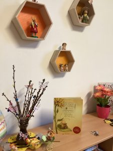 Ein Bücherregal mit einer Pflanze, einem gelben Buch und verschiedenen Spielsachen. Darüber stehen drei sechseckige Wandregale mit kleinen Figuren und Tierdekorationen vor einer cremefarbenen Wand.