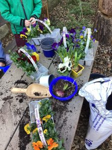 Kinder pflanzen bunte Blumen in recycelten Behältern auf einem Holztisch im Freien. Auf dem Tisch sind Erde, Blumentöpfe, Gartengeräte und ein teilweise geöffneter Sack mit Kompost zu sehen.