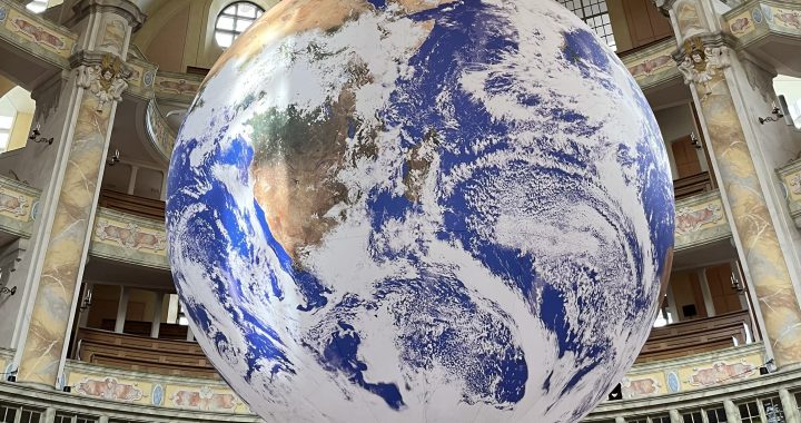 Ein großer, detaillierter Globus, der die Erde abbildet, hängt in einem prächtigen, verzierten Gebäude mit Bögen, Säulen und aufwändigen Verzierungen. Auf dem Globus sind vor allem Nord- und Südamerika abgebildet.