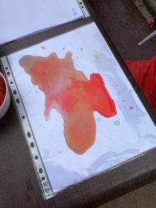 Ein Blatt Papier in einer Plastikhülle zeigt einen bunten Flüssigkeitsfleck in Rot-, Orange- und Gelbtönen, der einer abstrakten Form ähnelt. Das Papier liegt auf einem dunklen Tisch, daneben stehen eine Schüssel und ein roter Stoff.