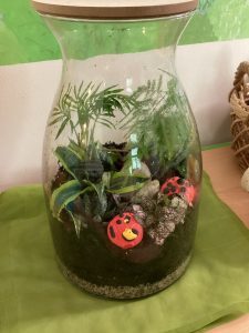 Ein Terrarium aus einem Glasgefäß mit grünen Pflanzen, Erde, Steinen und zwei roten Marienkäfer-Dekorationen darin, das auf einem grünen Tuch steht, daneben ein Weidenkorb.