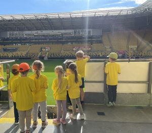 Eine Gruppe von Kindern in gelben Trikots und Mützen steht an einem sonnigen Tag in einem Stadion, den Blick auf das Spielfeld gerichtet und mit dem Rücken zur Kamera.