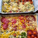 Zwei rechteckige Tabletts mit hausgemachter Pizza, perfekt für die Kita Knirpse, sind mit Käse, Peperoni, Kirschtomaten, Mais, grünen Erbsen und gemischtem Gemüse belegt - jedes Teil mit einer einzigartigen Kombination von Belägen.