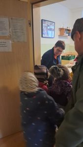 Eine Gruppe von Kindern mit Wintermützen und Mänteln betritt einen Raum, in dem ein Erwachsener an einem Schreibtisch mit Kisten sitzt; ein weiterer Erwachsener steht in der Nähe. In dem Raum gibt es ein Regal, Fotos an der Wand und Zettel an der Tür.