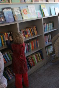 Ein kleines Kind in einem rot-schwarz karierten Kleid betrachtet bunte Bücher in einem niedrigen Bibliotheksregal und streckt beide Hände in die Höhe. Die Regale sind mit Kinderbüchern gefüllt, und andere Personen sind teilweise in der Nähe zu sehen.