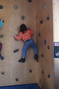 Ein kleines Kind in einem leuchtend orangefarbenen Pullover und Jeans klettert an einer Indoor-Kletterwand und hält sich mit Händen und Füßen an bunten Griffen fest.