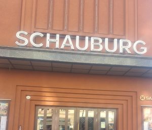 Eingang eines Gebäudes mit einer großen Leuchtreklame mit der Aufschrift „SCHAUBURG“ über den Glastüren. Das Gebäude ist braun und mit dekorativen vertikalen Linien verziert; zu beiden Seiten des Eingangs sind Plakate angebracht.