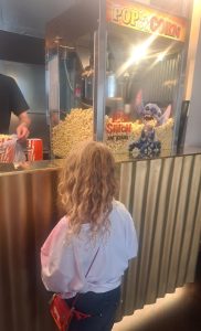 Ein kleines Kind mit lockigem blondem Haar steht an einer Verkaufsstheke und blickt auf eine Popcornmaschine, die mit einem „Lilo & Stitch“-Schild verziert ist und in der sich eine Stitch-Plüschpuppe zwischen dem Popcorn befindet. Im Hintergrund serviert ein Erwachsener Popcorn.
