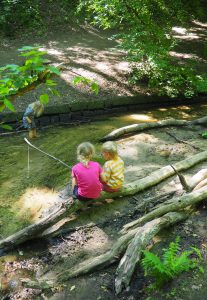 Drei Kinder spielen an einem seichten, sonnenbeschienenen Bach in einem Waldgebiet. Zwei sitzen auf großen Baumwurzeln am Wasser und halten sich an Stöcken fest, während das dritte im Bach steht und nach unten schaut. Üppiges grünes Laub umgibt sie.